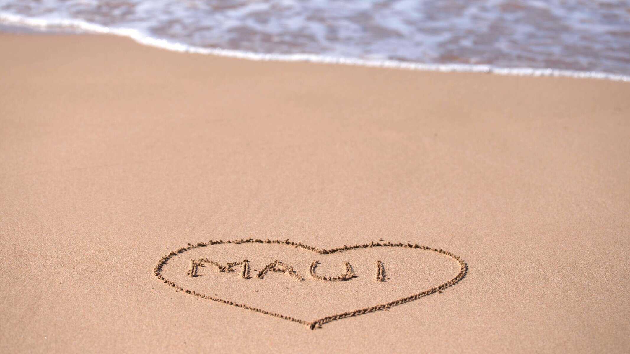 The word 'Maui' is written in the sand in a heart shape on Maui beach with waves.