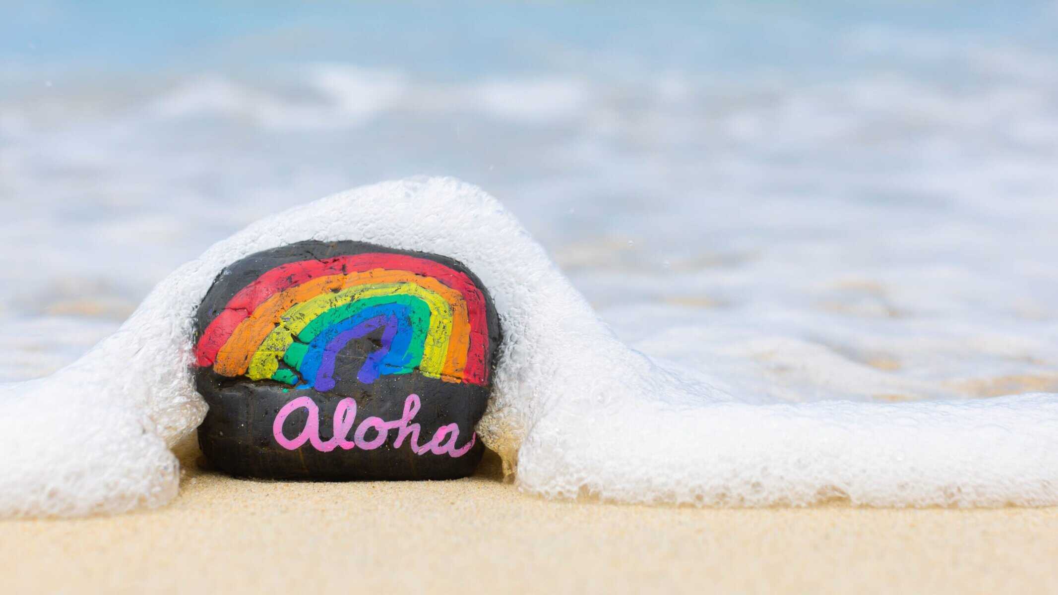 Aloha inscribed on a rock with rainbow hues, set against Maui's white sand beach.
