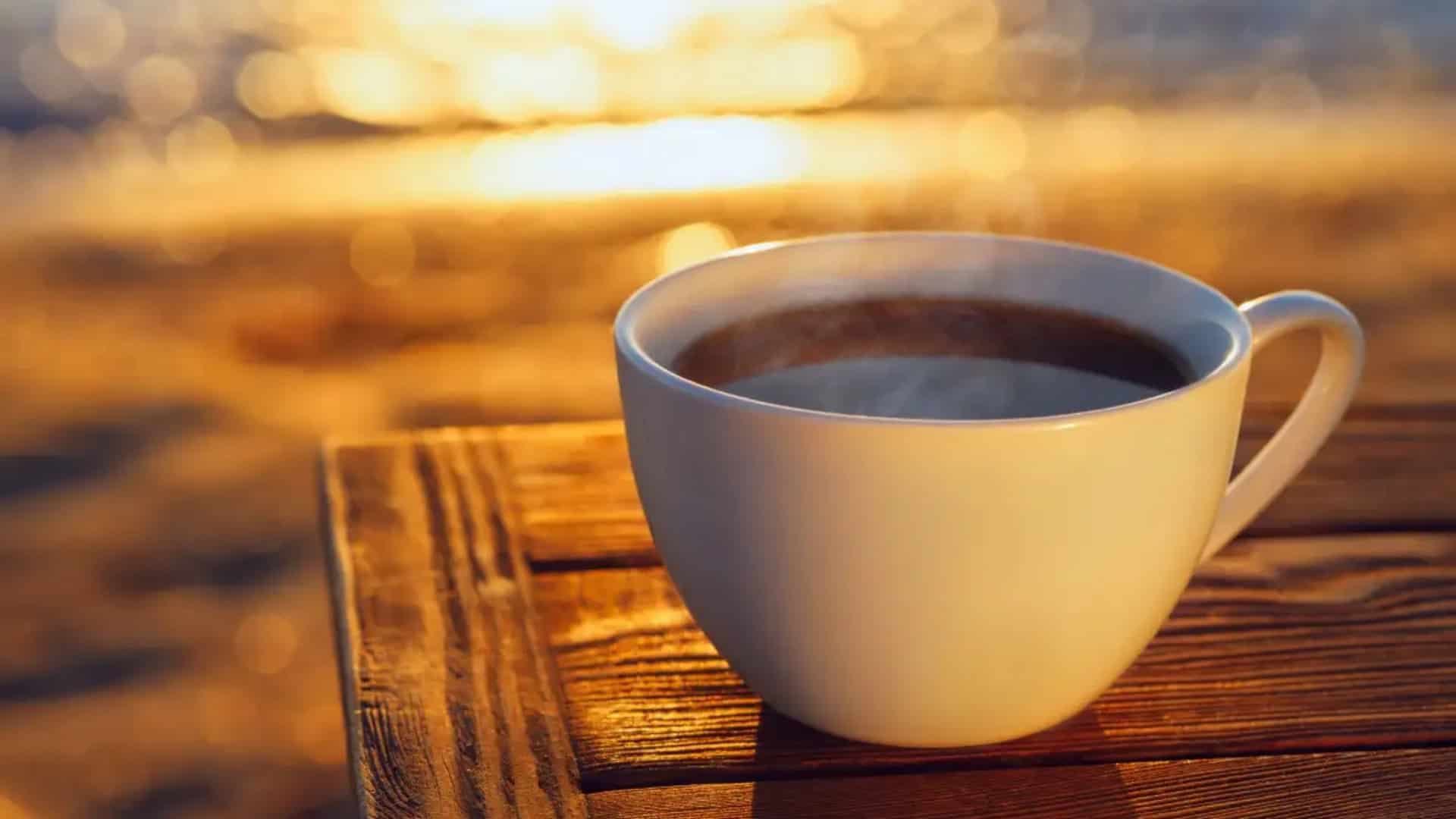 A steaming cup of black coffee sits on the sandy beach in Maui, framed by a stunning sunset over the ocean.