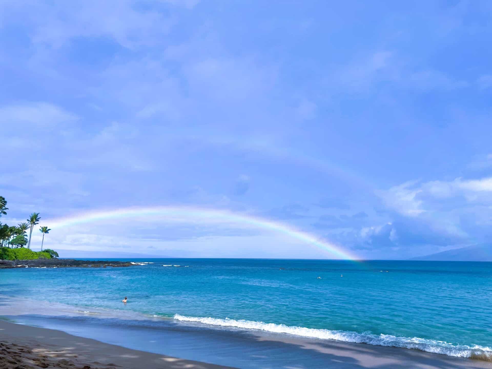A scenic view of a Maui beach under a rainbow, highlighting the beauty of rain and sunshine together.